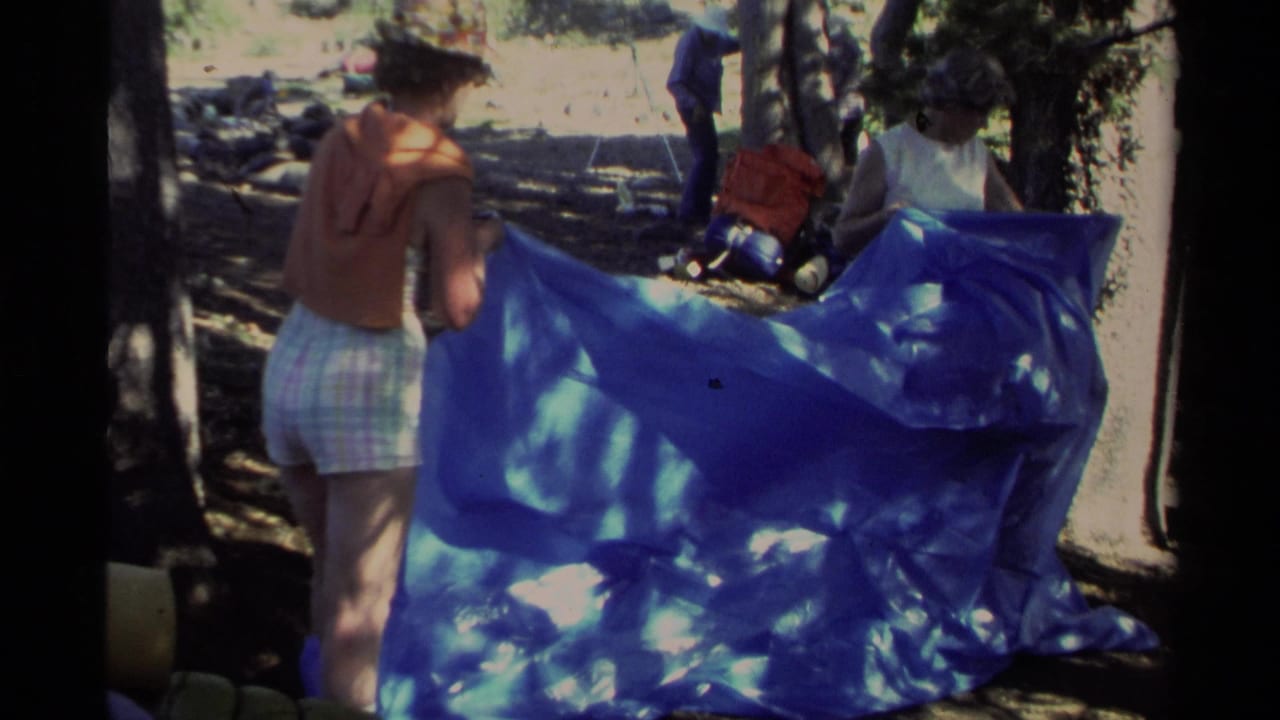 Adults at Lake Tahoe appear to adjust a blue tarp near tree trunks, with camping bags in the background.