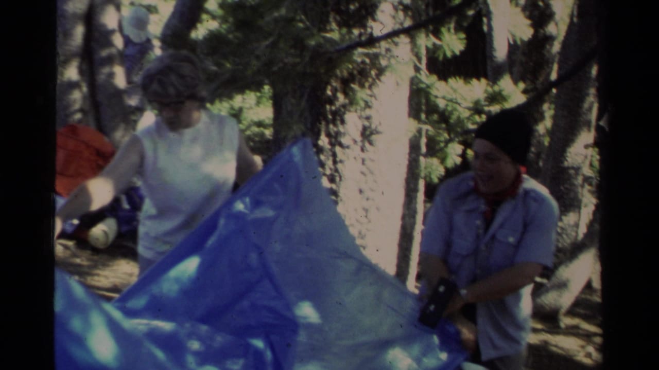 1978 Lake Tahoe home-movie frame showing two adults setting up a blue tarp among trees near a campsite.