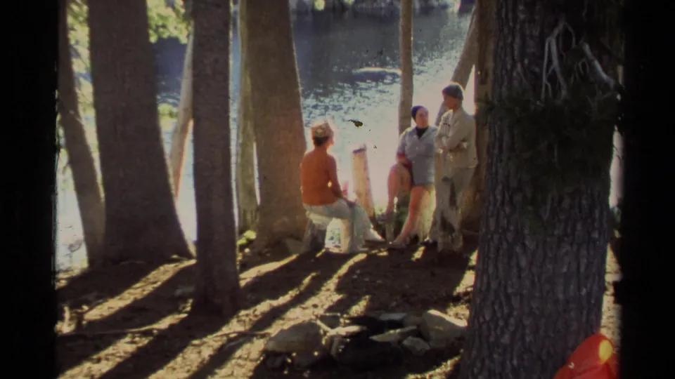 1978 Lake Tahoe frame of adults relaxing and talking beside the water under tall shoreline trees.