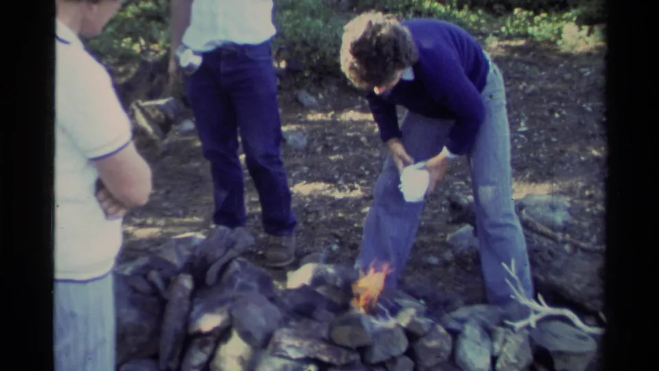 At a Lake Tahoe campsite, a man tends a small flame in a stone fire ring as other men stand near him.