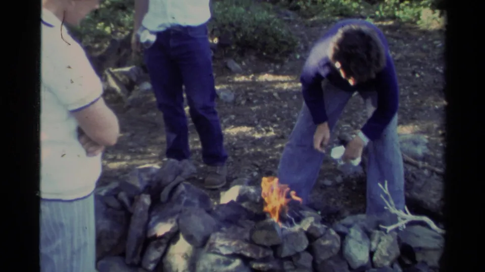 A man in a blue sweater tends a small campfire in a stone ring at a wooded Lake Tahoe campsite.