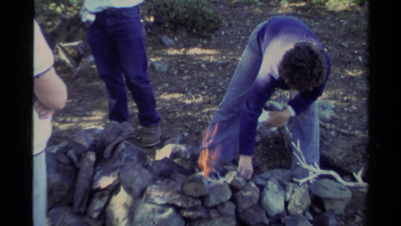 At Lake Tahoe in 1978, a young man in a purple sweater tends a small fire inside a stone ring as another person stands close by.
