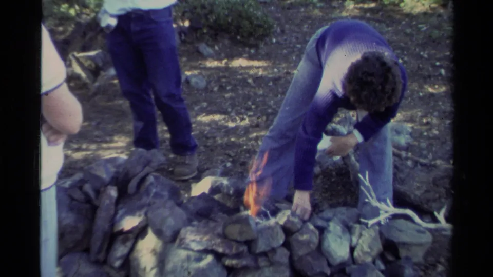 At Lake Tahoe in 1978, a young man in a purple sweater tends a small fire inside a stone ring as another person stands close by.