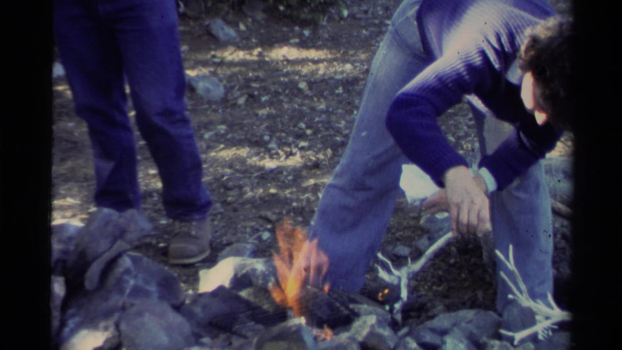 Man leans toward flames in a stone fire pit with another person's legs visible behind him.