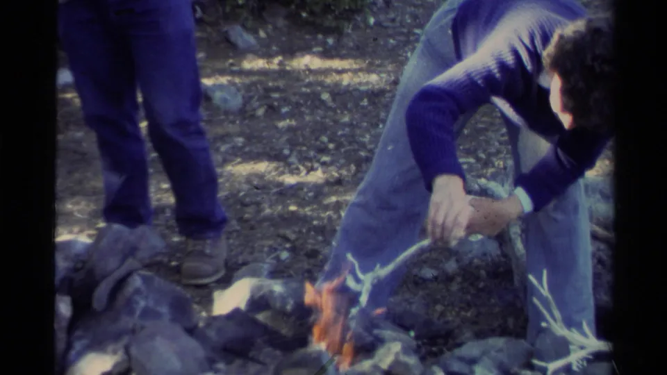 The home-movie frame shows a closer view of the man reaching toward the small fire, with rocks, branches, jeans, and forest floor visible.