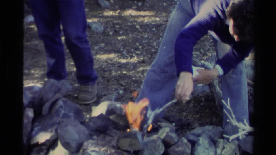At the end of the clip, the man remains crouched near the Lake Tahoe campfire, adjusting branches within the rock-lined pit.