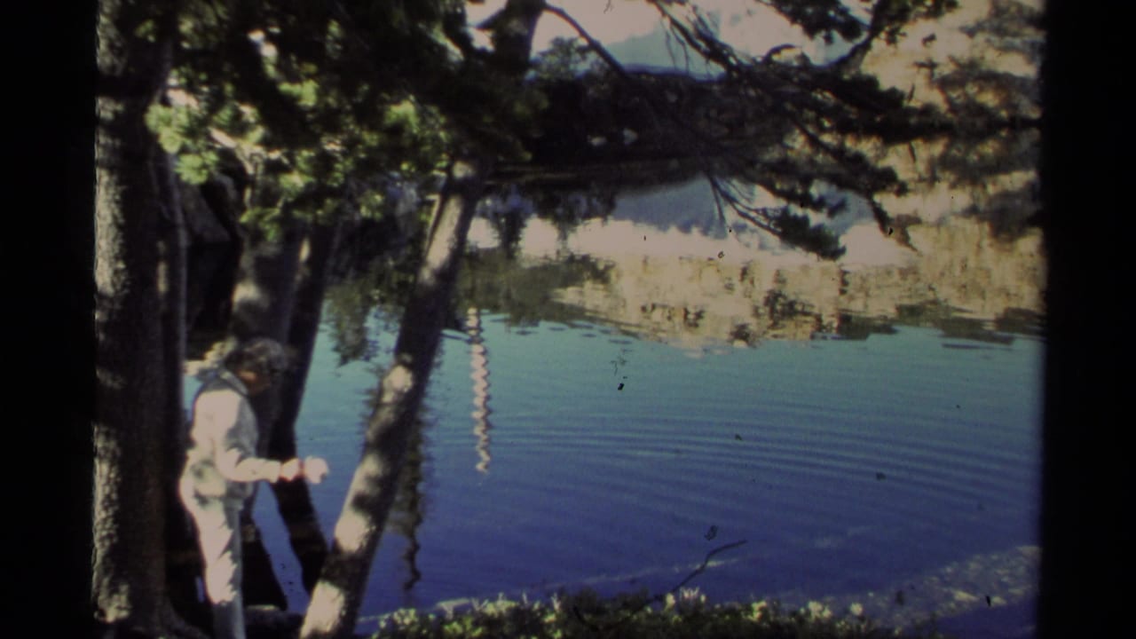 Lake Tahoe home-movie frame showing a person near pine trees at the water's edge, with blue lake water and distant reflected terrain.