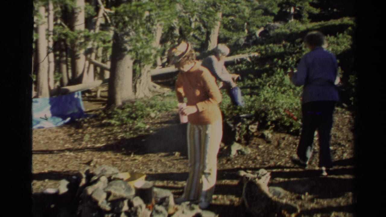 Women stand around a wooded campsite with a stone fire pit and lake visible nearby.