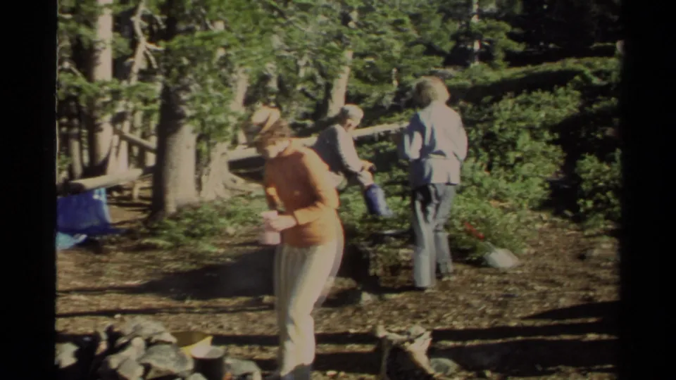 1978 Lake Tahoe camping frame showing women around a shaded fire pit area, with a blue tarp and forest floor visible.