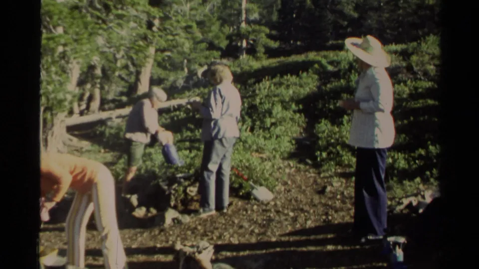 1978 Lake Tahoe home-movie frame of women gathered at a forest campsite, with a stone fire pit, pine trees, and camping gear nearby.