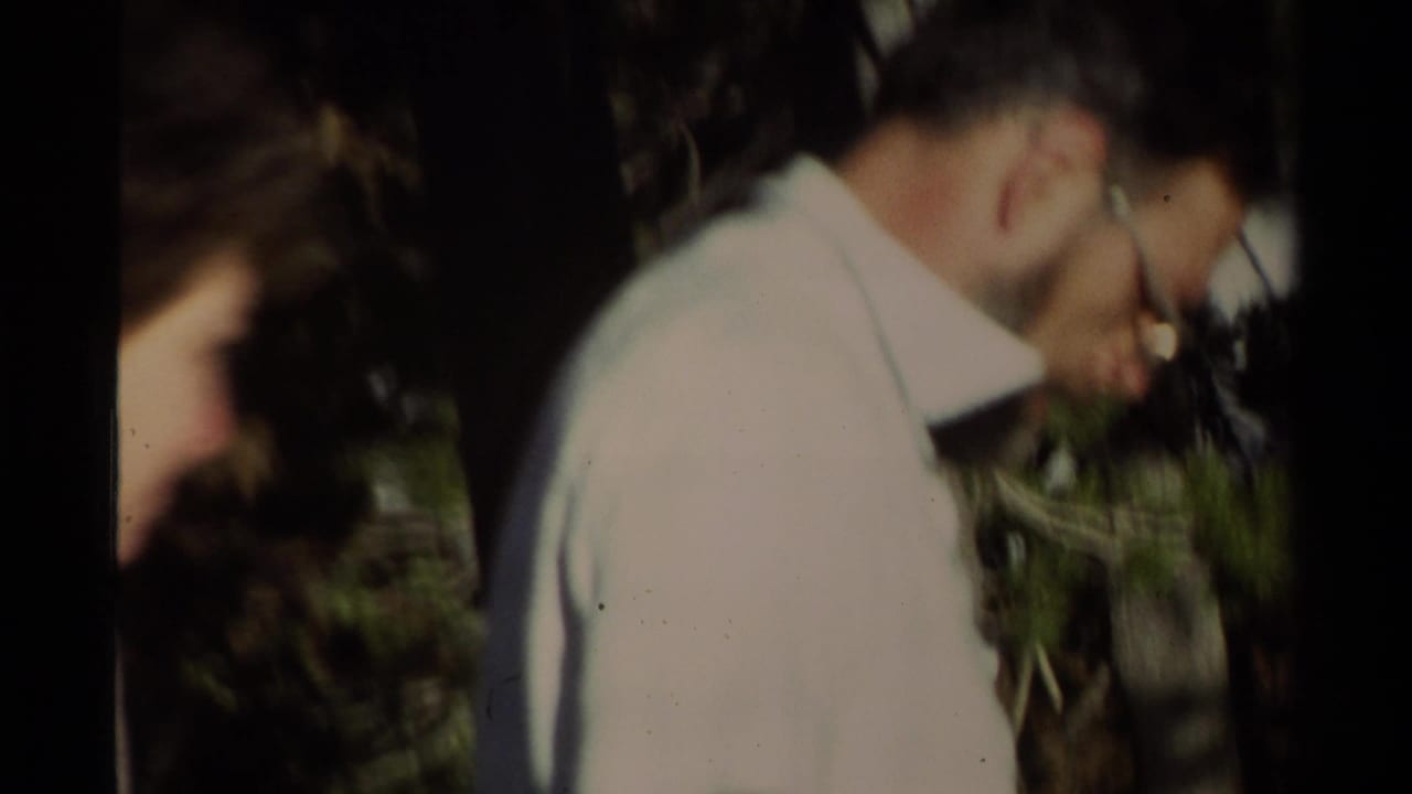 Soft 8mm close-up of an adult man in glasses and a light shirt moving through a shaded forest campsite area.
