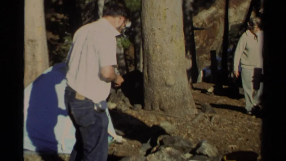 1978 Lake Tahoe campsite frame with a man near trees, rocks, and a tent or tarp while another adult stands nearby in shade.