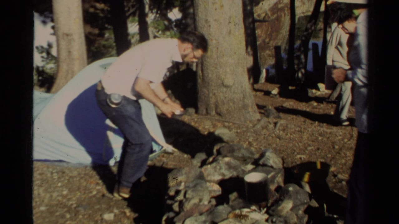 Man in a white shirt bends over a stone fire pit beside a blue tent in a wooded campsite.