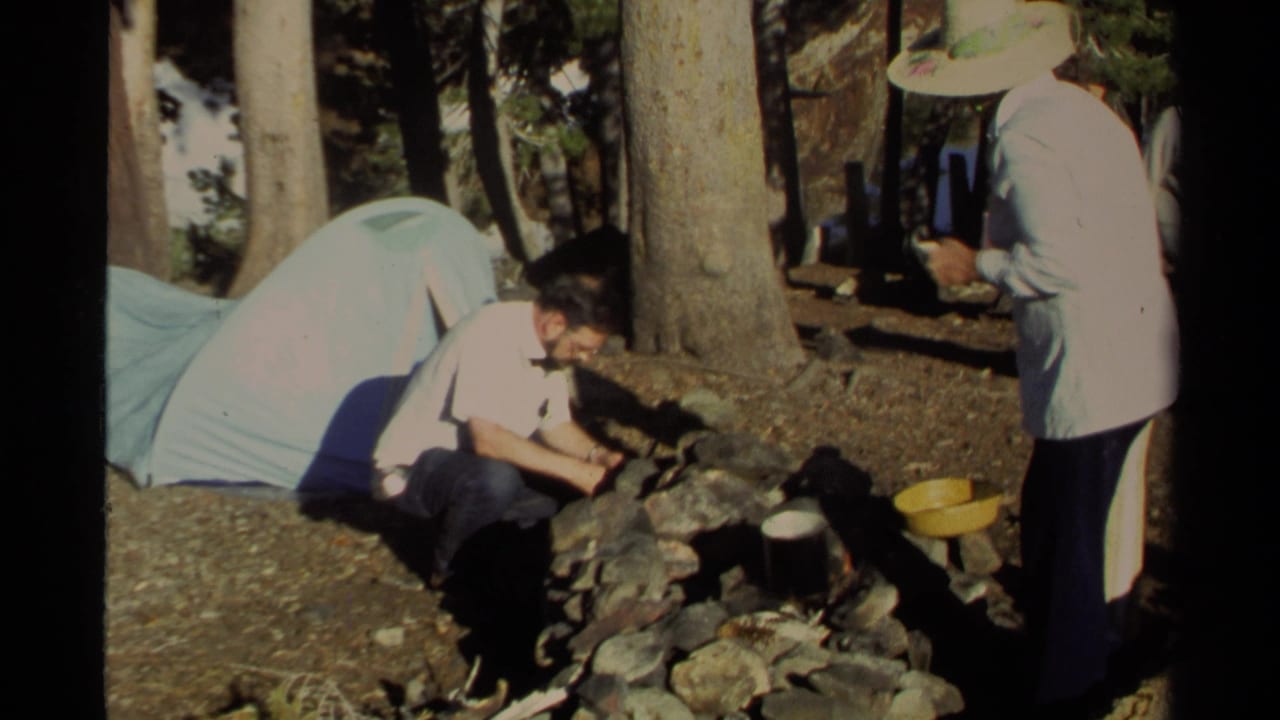 At the wooded Lake Tahoe campsite, a crouching man tends a stone fire ring as a woman in a wide-brimmed hat watches beside the tent.