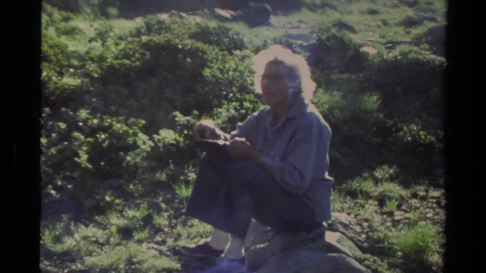 An older woman sits on a rock in bright Lake Tahoe greenery, holding a small object during the 1978 home-movie camping scene.