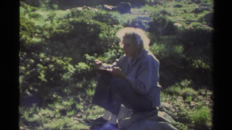 The 1978 Lake Tahoe home-movie footage shows an older woman seated on a rock, looking down at something she holds in the wooded campsite area.