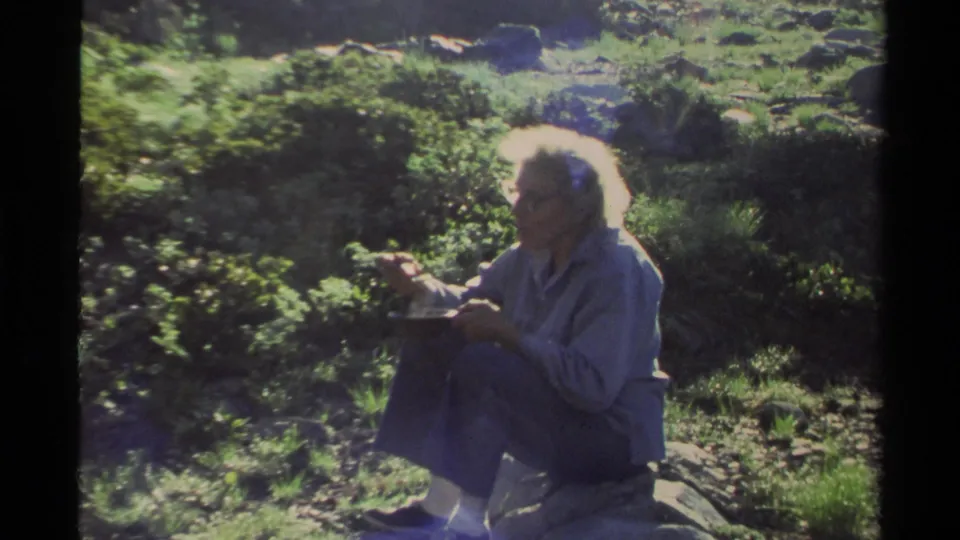 Adult woman with white hair and glasses sits on a rock eating from a plate in 1978 Lake Tahoe home-movie footage.