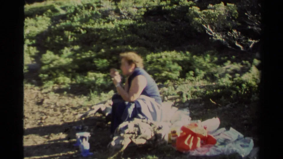 Adult brown-haired woman sits on a rocky patch eating beside cups, bags, and fabric in 1978 Lake Tahoe footage.