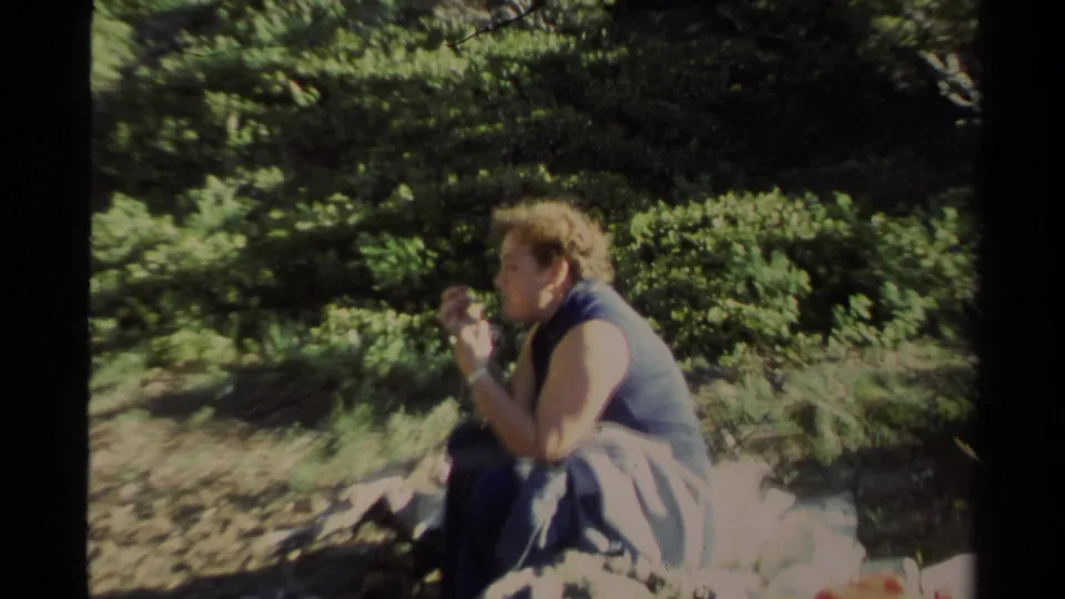 The brown-haired woman continues eating in a sunlit forested rocky area, filmed with soft handheld 8mm framing.