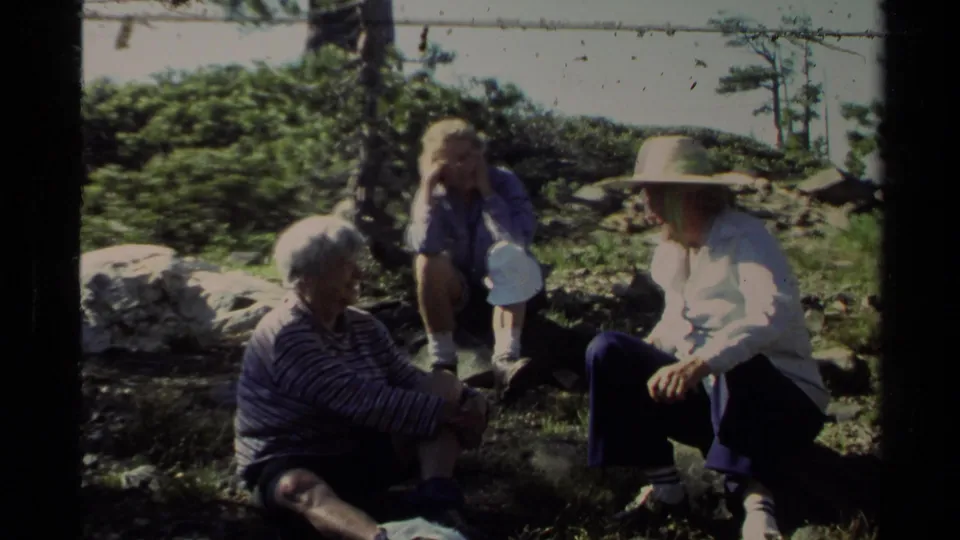Three adults rest together outdoors in a 1978 Lake Tahoe home-movie frame, with grass, rocks and trees around them.