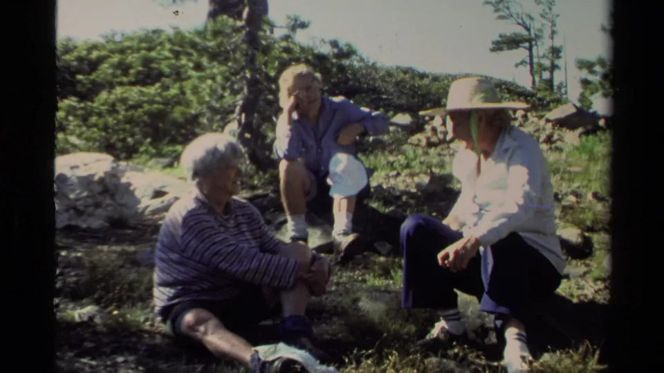 1978 Lake Tahoe home-movie frame showing adults and a child resting on rocky ground with shrubs and trees behind them.