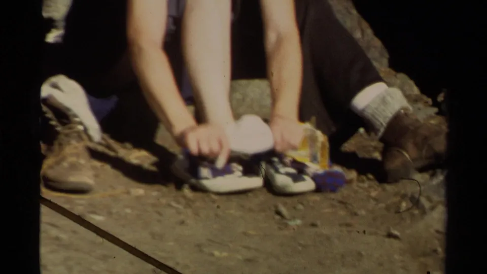 Close-up from the 1978 home movie showing a child handling sneakers on dirt ground near an adult's hiking boots.
