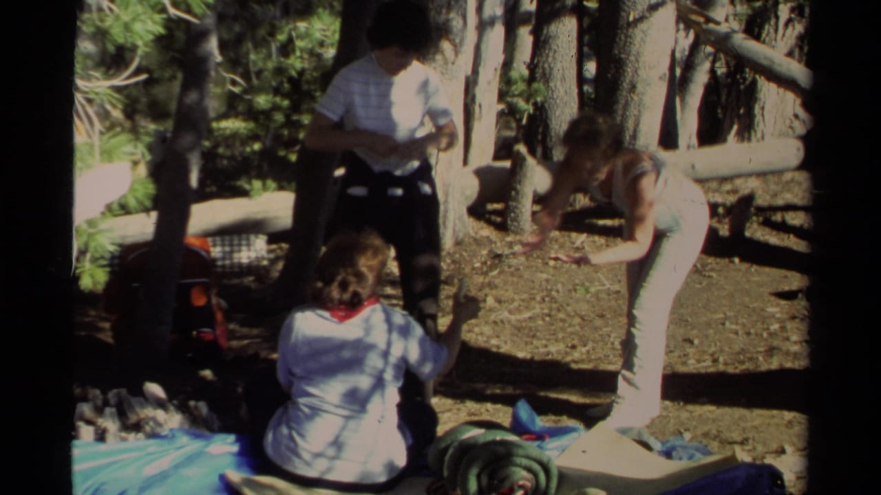 1978 Lake Tahoe home-movie frame showing three campers gathered near a tarp and sleeping gear in a sunlit forest clearing.