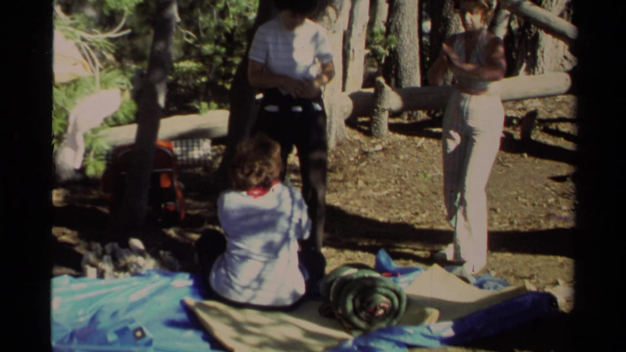 Campers stand and sit near camping gear on a tarp in a shaded forest clearing.