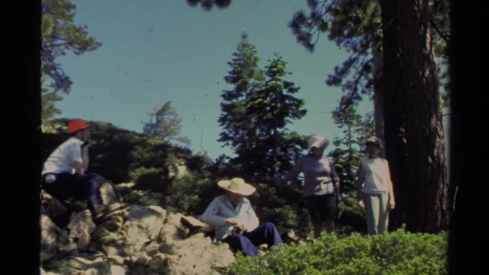 Several adults in the Lake Tahoe home-movie scene stand or sit on a rocky pine slope, with one figure gesturing upward.