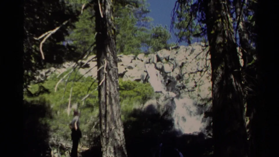 A distant hiker stands in a pine forest looking toward a rocky waterfall in the 1978 Lake Tahoe home movie.