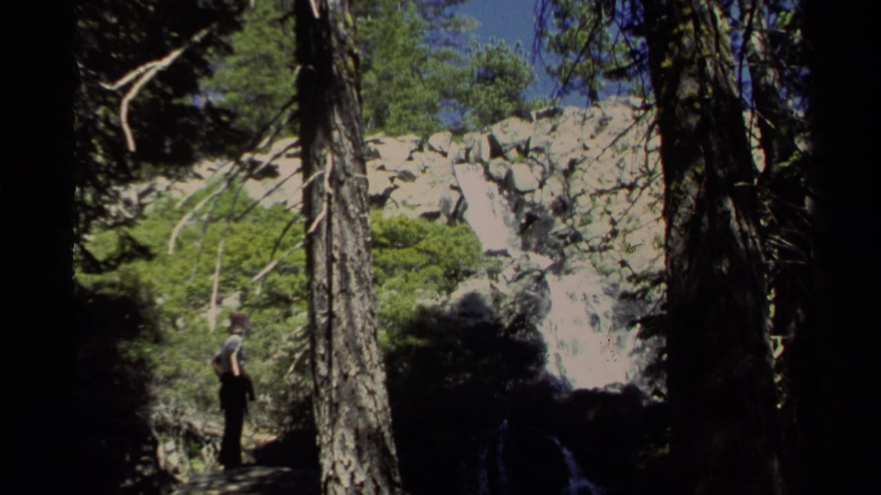 1978 Lake Tahoe 8mm frame showing a woman in a forest clearing near pine trunks, sunlit rocks, and a shadowed waterfall.