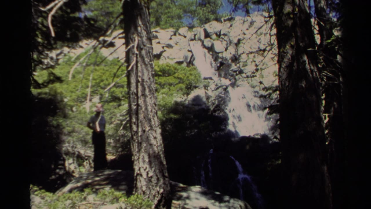 1978 Lake Tahoe 8mm frame with a woman near a waterfall, raising her arms amid pine trees, rocks, and bright green foliage.
