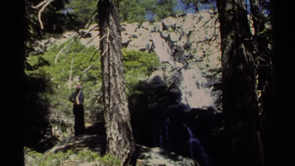 1978 Lake Tahoe 8mm frame with a woman near a waterfall, raising her arms amid pine trees, rocks, and bright green foliage.
