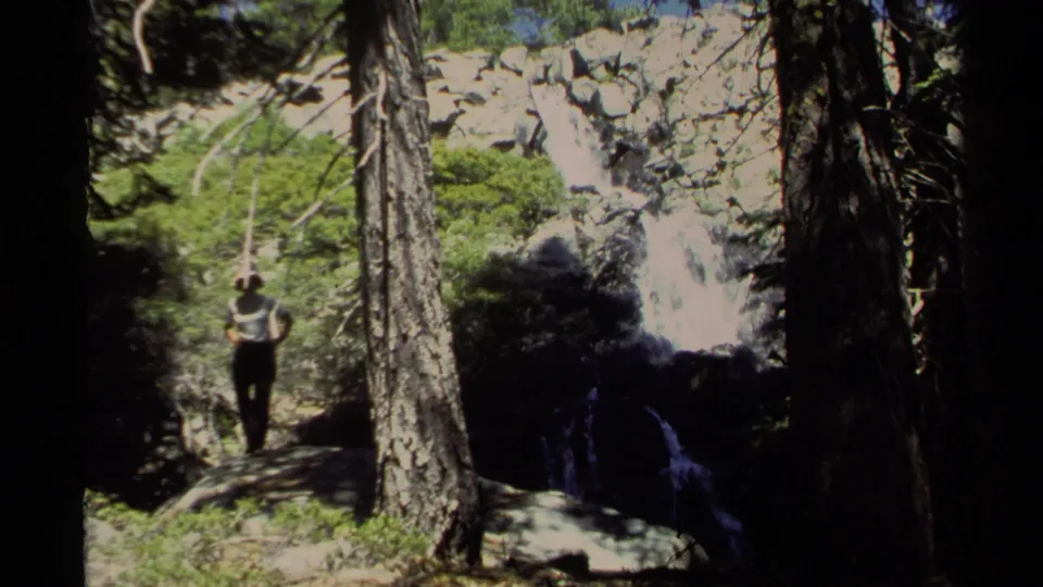 1978 Lake Tahoe 8mm frame showing the woman closer to camera on a shaded forest ledge with pine trunks, rocks, foliage, and falling water.