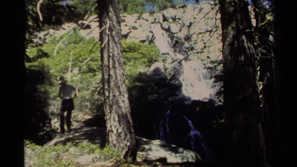 1978 Lake Tahoe 8mm frame showing a woman from behind looking toward a waterfall, with pine trunks, rocks, and bright foliage around her.