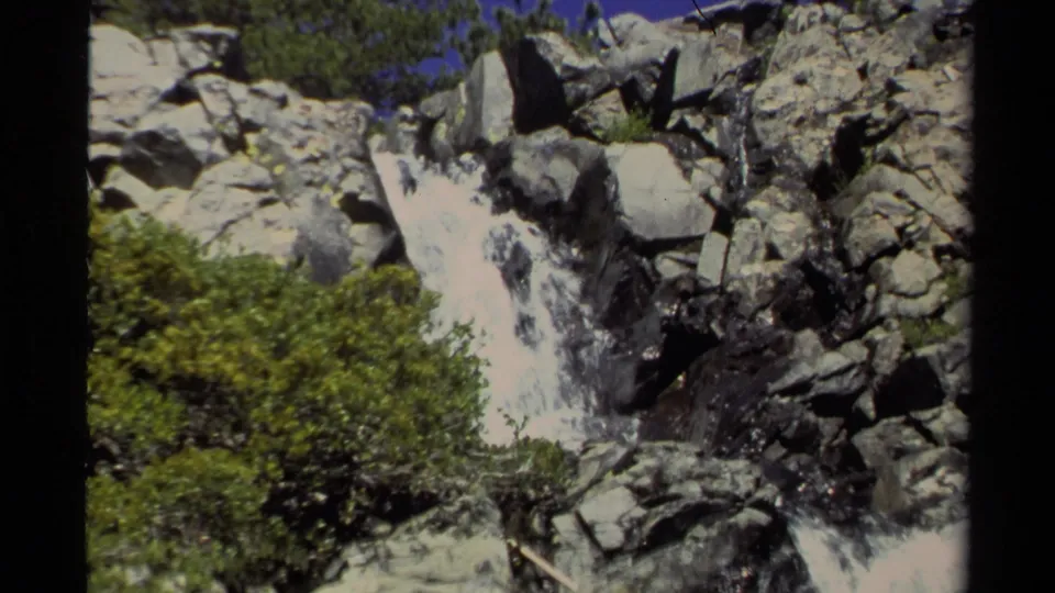 1978 Lake Tahoe home-movie frame showing a rocky waterfall, brush and a distant person standing above the rushing water.