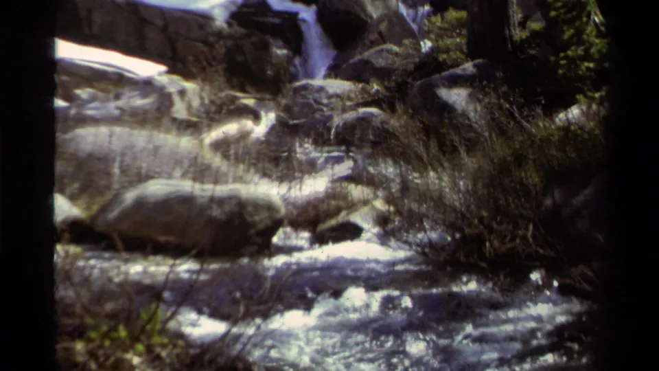 Lake Tahoe creek scene from 1978 8mm footage, with rippling water, rocks, brush, and a small cascade behind.