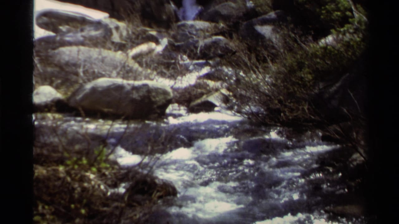 1978 Lake Tahoe 8mm frame showing a rocky stream with foamy water, shrubs, and sunlit boulders along the bank.