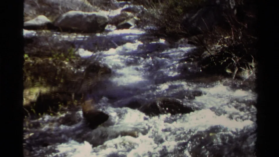Lake Tahoe home-movie frame with bright rapids moving over dark rocks, framed by brush and rounded boulders.