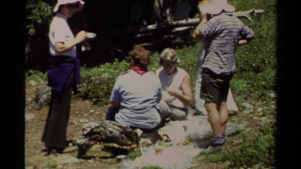 Several adults picnic in a grassy Lake Tahoe area, with hats, a blanket, rocks, and food items visible.