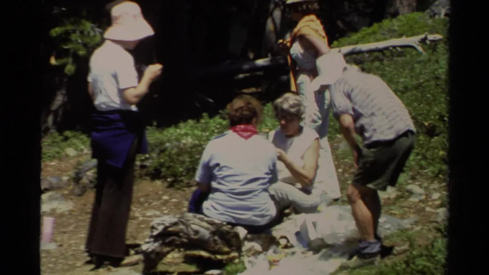 Adults in summer clothing gather around a Lake Tahoe picnic blanket, one leaning in while others sit on rocks.