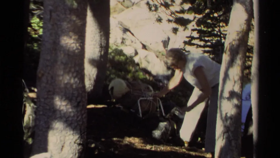 A person in a light sleeveless top leans over camping gear in the shaded 1978 Lake Tahoe forest scene.