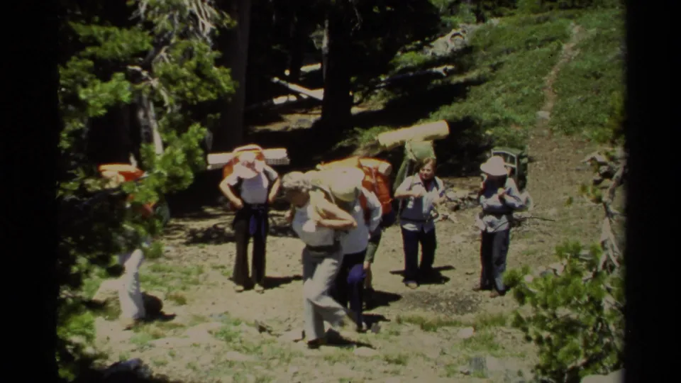 1978 Lake Tahoe home-movie frame of adult hikers with external-frame backpacks standing on a forest trail in bright sunlight.