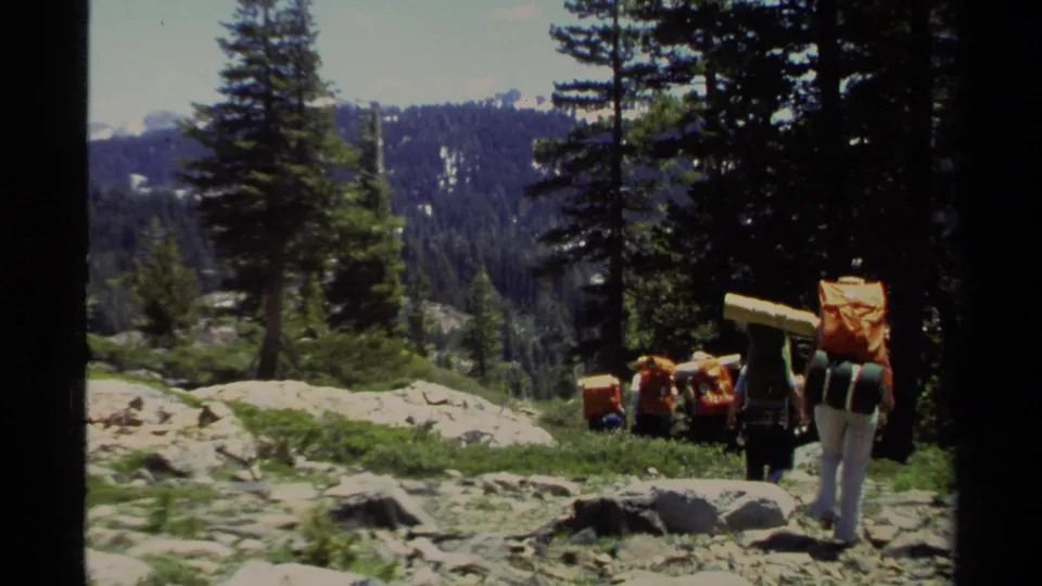 1978 Lake Tahoe home-movie frame showing hikers with large packs walking downhill through a rocky pine forest trail.