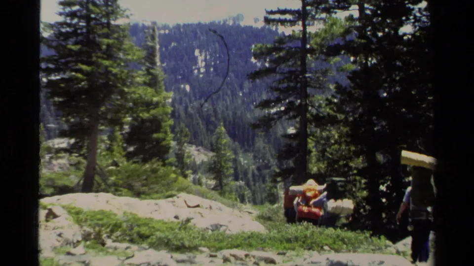 1978 Lake Tahoe footage frame with backpackers moving away through pines, rocky ground, and distant wooded slopes.