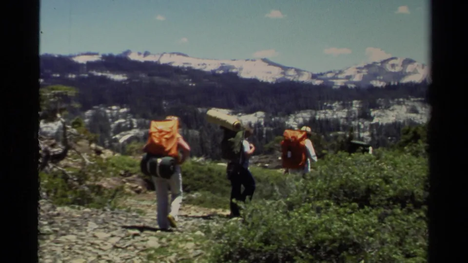 Lake Tahoe home-movie frame showing hikers with large packs on a rocky path below distant snowy mountains.