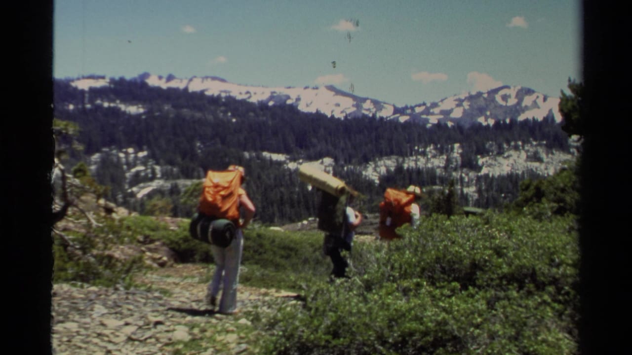 1978 Lake Tahoe home-movie frame of backpackers moving along a rocky trail with shrubs, pines, and snow-covered peaks beyond.