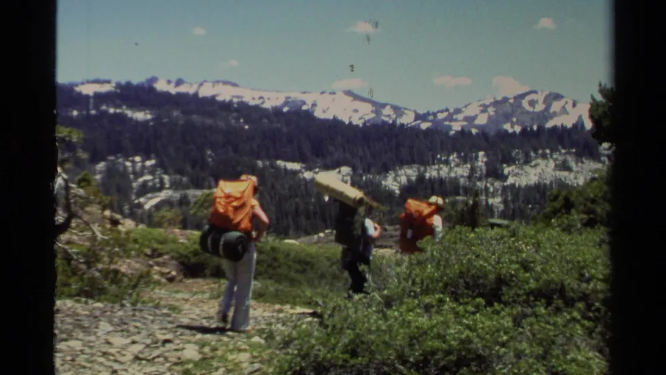1978 Lake Tahoe home-movie frame of backpackers moving along a rocky trail with shrubs, pines, and snow-covered peaks beyond.