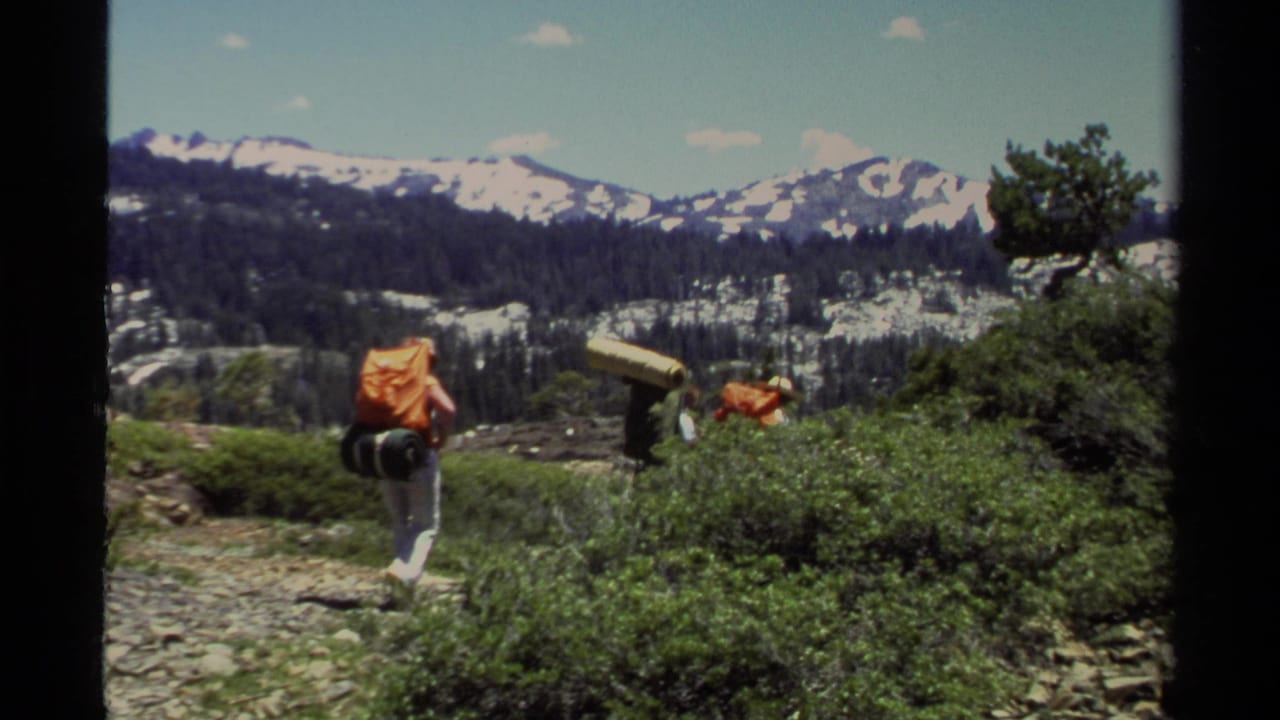 A Lake Tahoe backpacker in 1978 walks away from the camera on an alpine trail, with other hikers partly visible near shrubs.