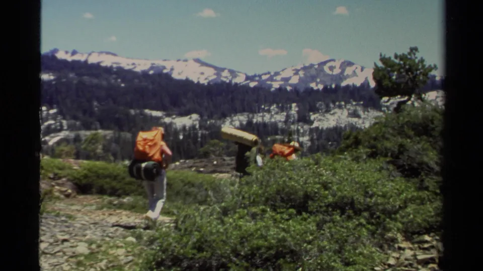 A Lake Tahoe backpacker in 1978 walks away from the camera on an alpine trail, with other hikers partly visible near shrubs.